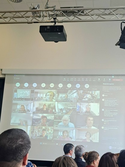 Foto aus einem Konferenz Raum mit Personen im Raum und auf der großen Leinwand viele Online Videos von Teilhabenden 
