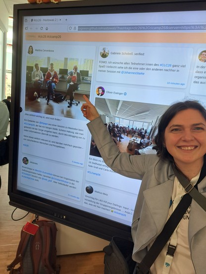 Freudige Frau zeigt auf die Mastowall weil dort ihr Beitrag im Mastodon steht